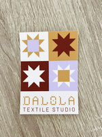 Sticker with quilt pattern design on a wooden surface, featuring the brand 'DALOLA Textile Studio'.