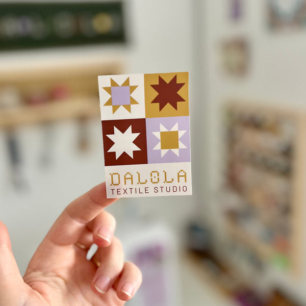 Hand holding a sticker with a pattern and 'DALOLA TEXTILE STUDIO' text against a blurred background.
