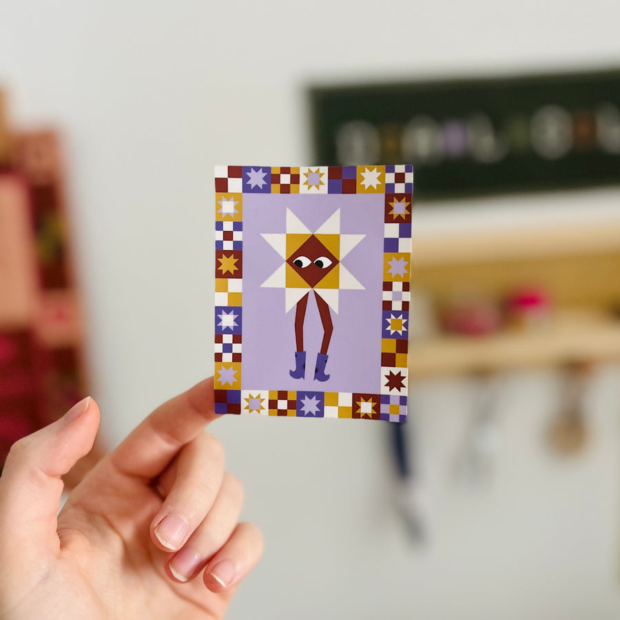 Hand holding sticker with quilt-motif character wearing cowboy boots against a blurred background.