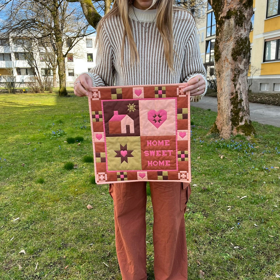 Person holding a quilt with 'Home Sweet Home' text in an outdoor setting