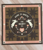 Decorative fabric panel with rabbits, heart, and text 'Home is where the heart is' on a geometric, quilted motif background.
