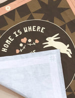 Decorative fabric panel with rabbits, heart, and text 'Home is where the heart is' on a geometric, quilted motif background.